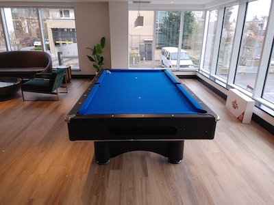 A pool table on a brown floor with a blue playing surface. There are windows on the walls, showing a white van outside.