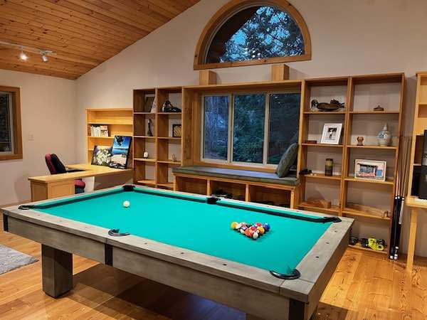 A green pool table in a room with wooden floors and walls and a tall, wooden shelf. 