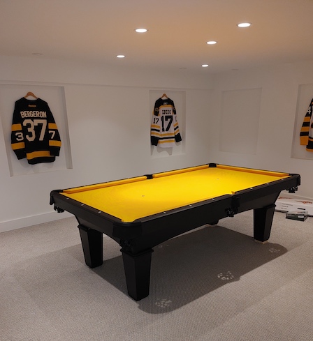 A yellow pool table in a carpeted room with hockey jerseys on the walls around it.