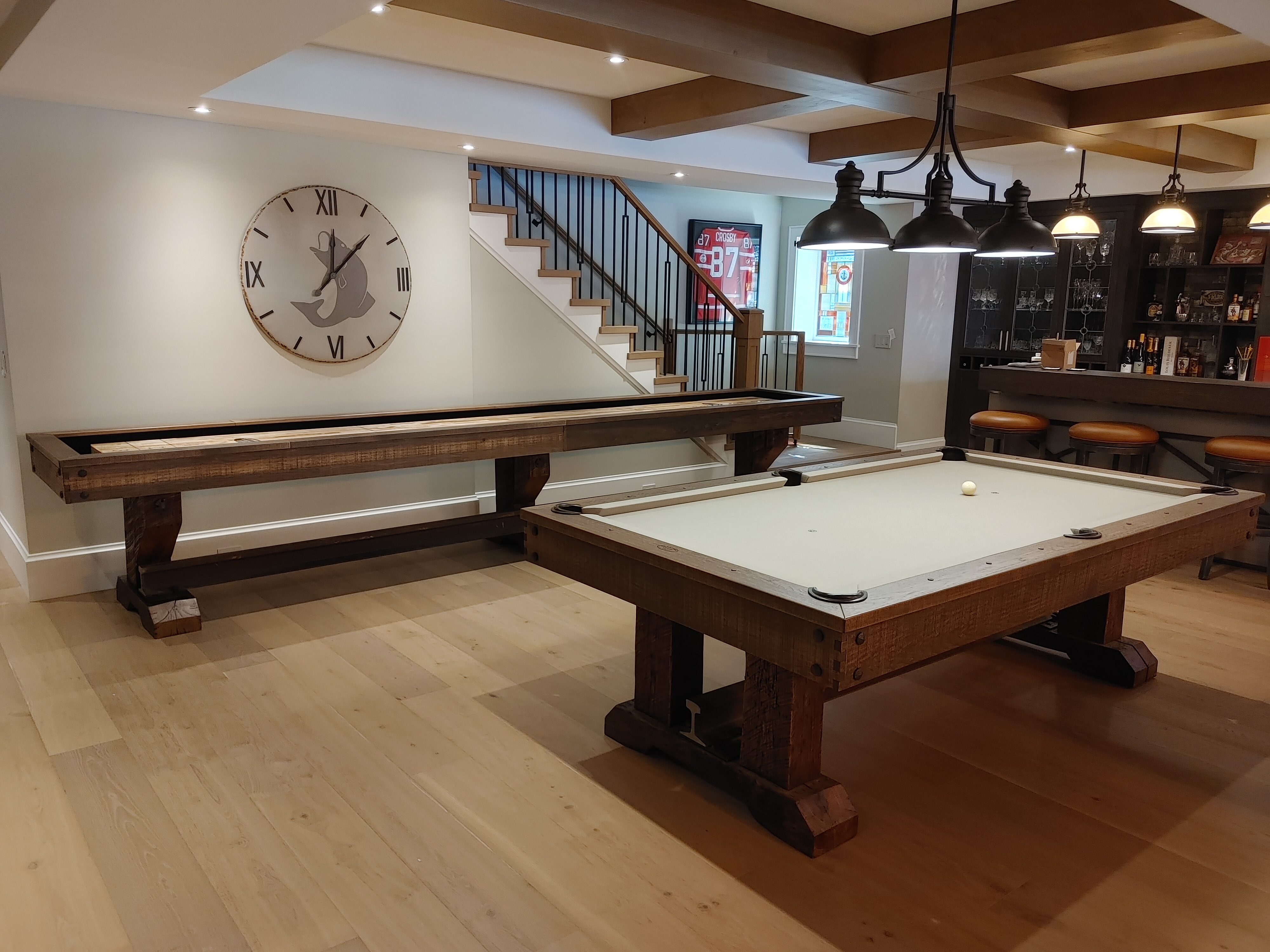 Pool table with light coloured felt in a rec room that has a shuffleboard table and bar in the background.