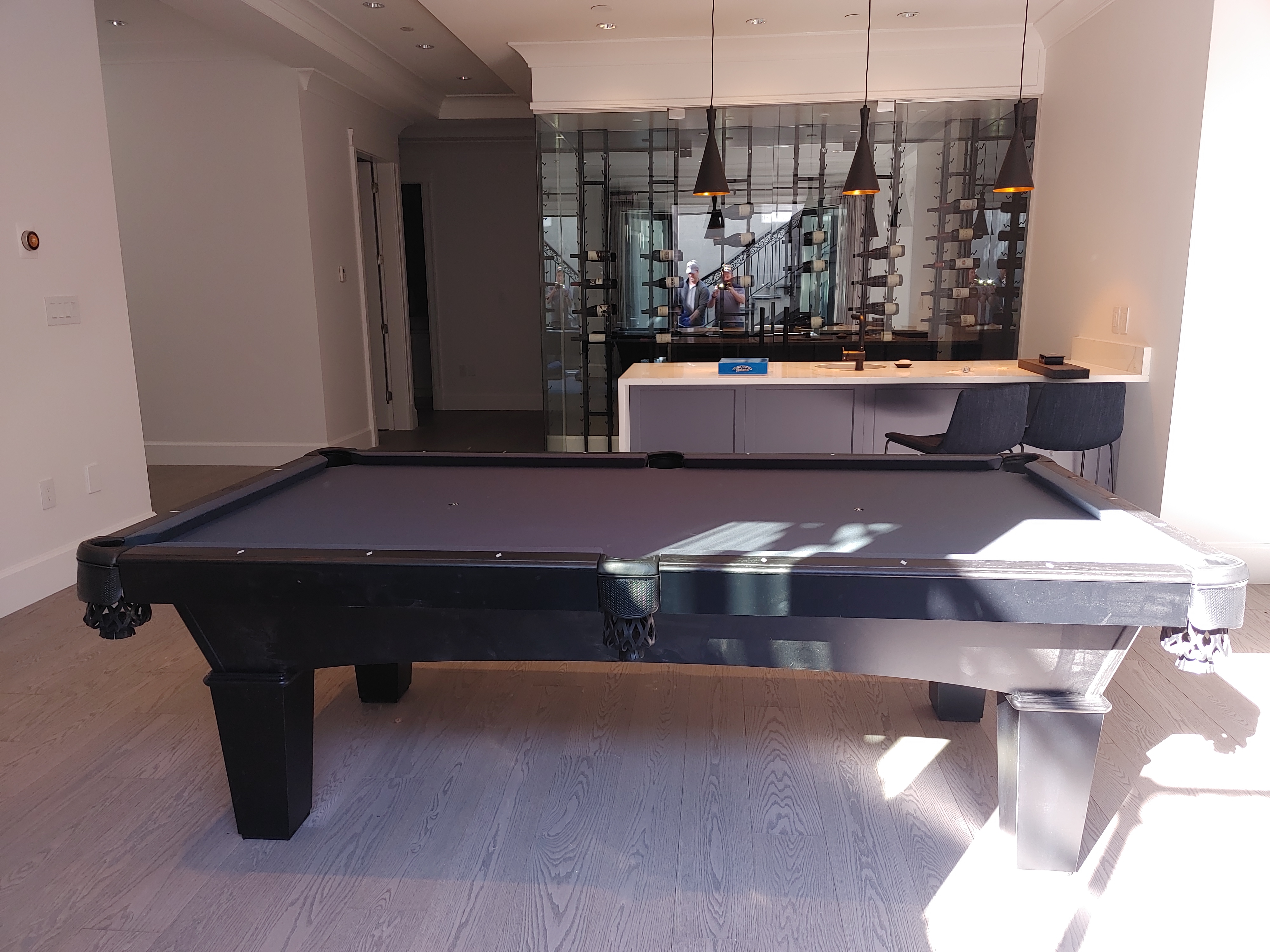 A pool table with dark cloth in a wood floor room with glass wine rack and bar in background.