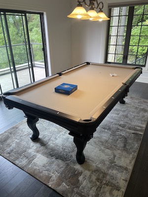 A pool table with brown bumpers and tan cloth under three hanging overhead lights