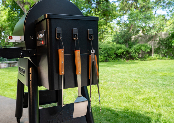 A Traeger wood pellet grill with three magnetic hooks on its side holding different grilling accessories.