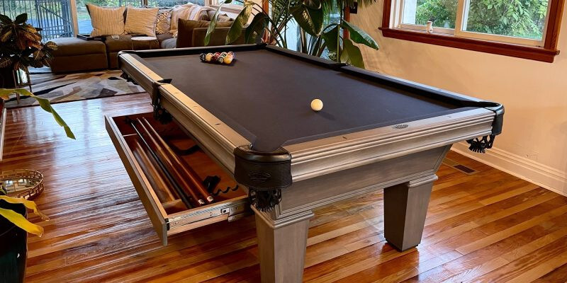 Modern pool table with black felt, built-in cue drawer storage, and hardwood floors in a bright living space