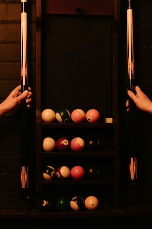 Billiard balls arranged on wall rack with hands holding two cue sticks under warm lighting.
