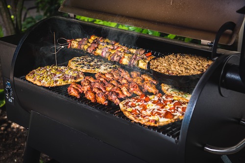 Alt description: An open barbeque with different foods cooking simultaneously, including vegetables, steak, and pizza.