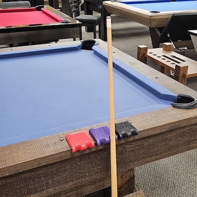 Blue-felt pool table with wooden cue stick and red, purple, and black Cue Claw cue holders on the rail.
