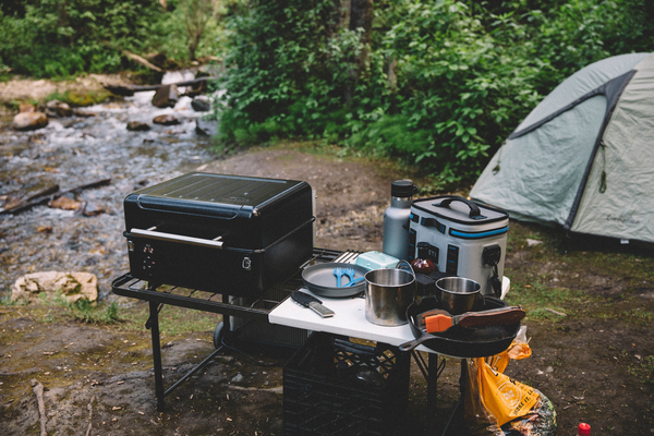 The Traeger Ranger wood pellet grill set up on a table at a campsite with a tent a river in the background.