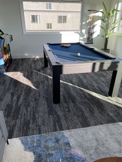 Pool table in a light filled room with game console and plants surrounding the table.
