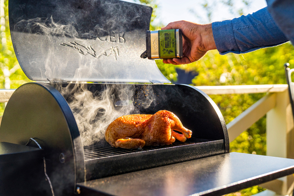 Man seasoning a whole chicken being cooked on a Traeger Tailgater wood pellet grill.