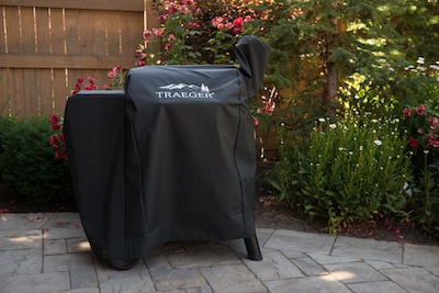 Traeger grill protected by a grill cover on a stone patio with wooden fence and rose flowers in the background.
