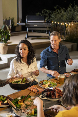 A group of friends enjoying a meal at night on a patio in the backyard with a Traeger Ironwood XL wood pellet grill in the background.