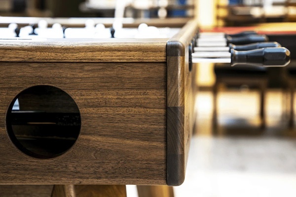 Close-up of a wooden foosball table with player rods and ball return opening in a recreation room.