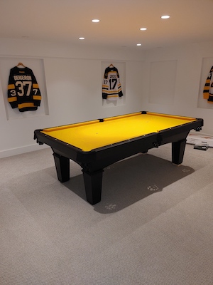 A pool table in a basement with picture frames on the surrounding walls, yellow hockey jerseys are inside the frames.
